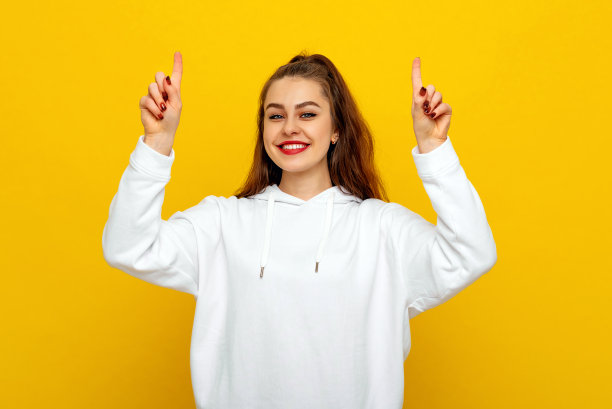 穿着白色休闲风格运动衫的开朗的黑发女孩大笑着，手指向上，展示着广告。室内工作室拍摄的黄色背景图片下载