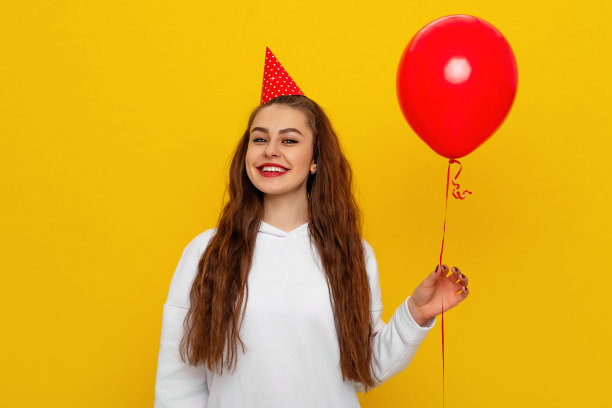年轻快乐的女人与黑色头发在休闲服装庆祝生日节日聚会举行红色空气膨胀的氦气球，孤立在平原黄色的背景工作室。假日与聚会的概念图片下载