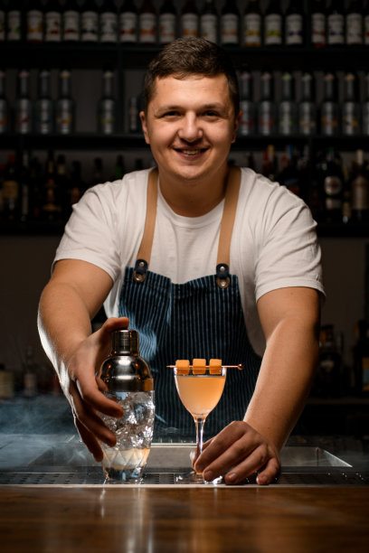年轻微笑的酒吧服务员专业地拿着透明的摇壶与冰块和一杯冷酒精鸡尾酒图片下载