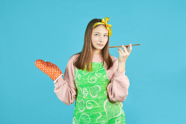 穿着厨房衣服的快乐女孩在蓝色背景上拿着木勺和锅架。穿围裙的年轻女子是一个有厨房用品的家庭主妇。图片下载