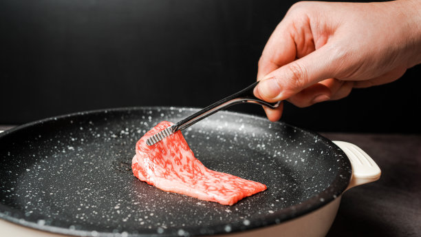 上等生牛里脊A5牛肉片，高大理石纹理，拿起，用烧烤钳在大理石涂层的不粘锅上烤，手放在黑色背景上。对于日式烧肉。图片下载