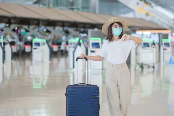 年轻女性戴着口罩带行李在机场行走，保护冠状病毒感染，亚洲女性旅行者戴着帽子。时间旅行后疫苗加强剂量的概念图片下载