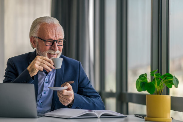 微笑的高级专业人员在办公室的办公桌上一边喝咖啡，一边使用笔记本电脑图片下载