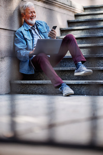 一位随意的年老商人坐在外面的楼梯上，用笔记本电脑工作。图片下载