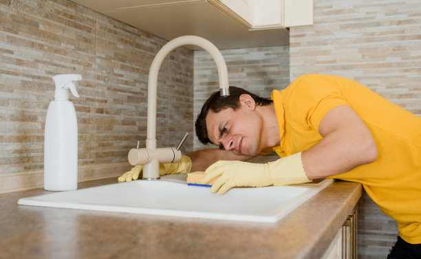 穿着黄色t恤、戴着黄色手套的年轻人正在用清洁喷雾和海绵清洁厨房的洗脸盆水槽。图片下载