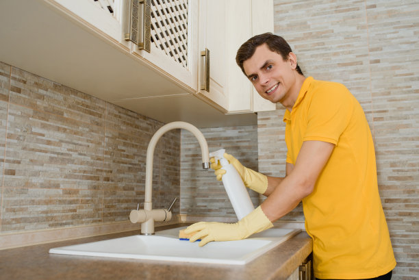 穿着黄色t恤、戴着黄色手套的年轻人正在用清洁喷雾和海绵清洁厨房的洗脸盆水槽。图片下载