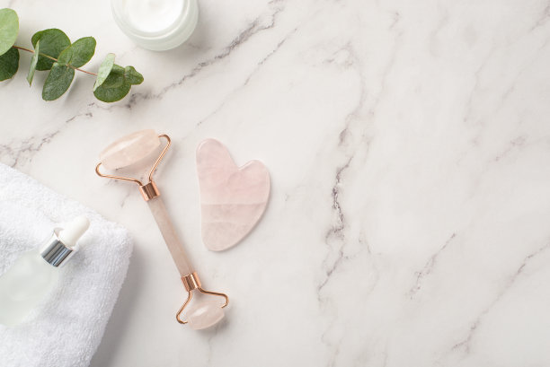 护肤概念。俯视图照片的玫瑰石英辊挂沙按摩膏罐玻璃透明瓶桉树和白色毛巾在白色大理石的背景与copyspace图片下载