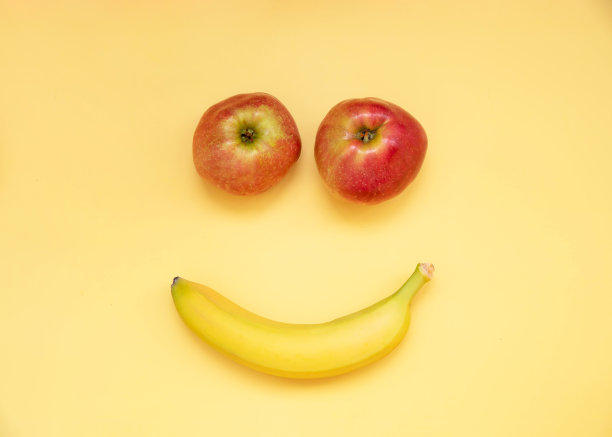 两个红苹果和一根黄香蕉躺在黄色的背景上，画着眼睛和微笑图片下载