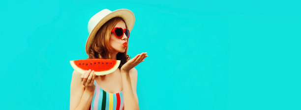 夏天的肖像美丽的年轻女子吹她的嘴唇与红色的口红发送空中吻与西瓜片戴着草帽在蓝色的背景，空白的拷贝空间的广告文字图片下载
