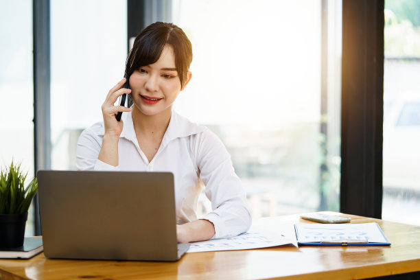 亚洲女企业家在办公室忙着工作。年轻的亚洲女性一边在办公桌上用电脑工作，一边用智能手机或手机聊天。图片下载