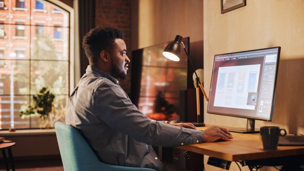 年轻英俊的非洲裔美国人在阳光时尚的阁楼公寓的桌面电脑从家里工作。有创意的男性查看社交媒体，浏览互联网。从大窗户看城市。图片下载