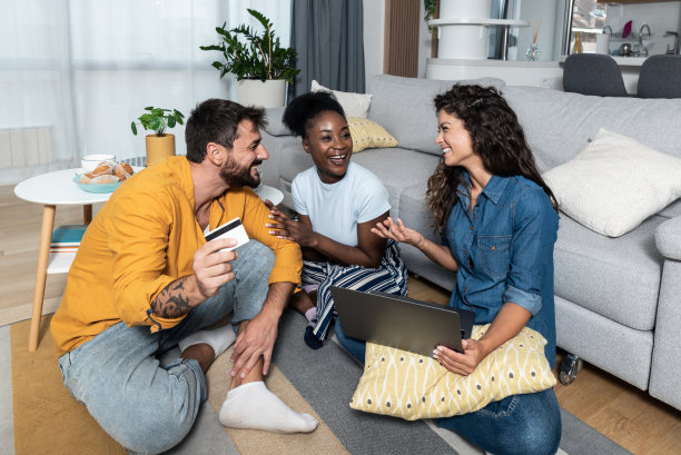 三个无忧无虑的年轻自由职业商业伙伴和老板，坐在家里的朋友们很高兴，因为他们刚刚尝试用信用卡在他们新的网上商店网站上购物。图片下载