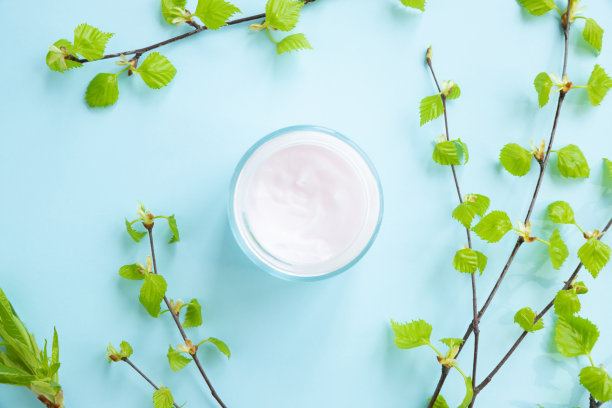 保湿霜在开放的玻璃罐中和白桦树枝与年轻的小叶子在蓝色的背景。平铺，俯视图。图片下载