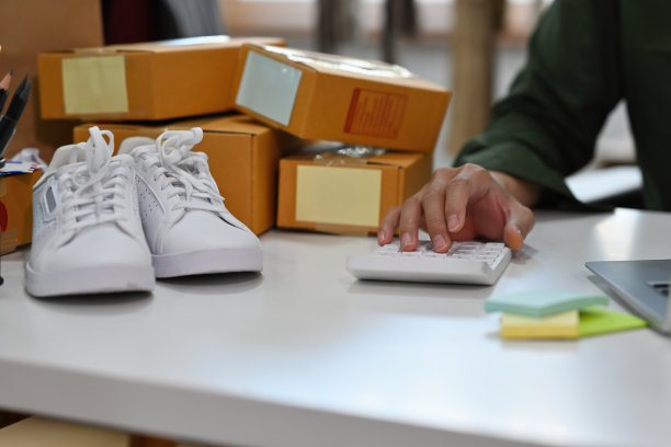 近距离观察一名上班族坐在办公室里，使用笔记本电脑和计算器，箱子堆在桌子上，白色运动鞋在一旁，为中小企业、创业、金融、电子商务和送货概念。图片下载