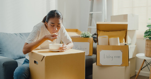 年轻的亚洲女人搬家坐在沙发上吃方便面后拆包。图片下载