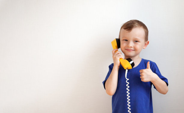 身穿蓝色t恤的4岁男孩正在白色背景的黄色复古手机上聊天。男孩用手做了个班级手势图片下载
