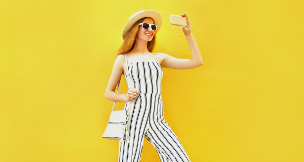 美丽微笑的女人的肖像通过智能手机自拍戴夏季草帽，白色条纹连体衣，手提包在黄色的背景图片下载