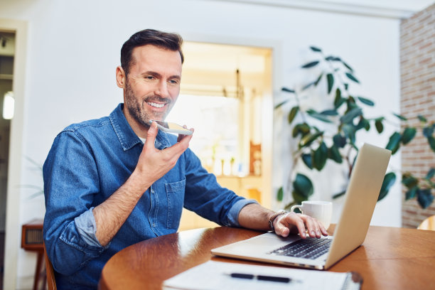 微笑的男人坐在餐桌上用手提电脑用扬声器打电话图片下载