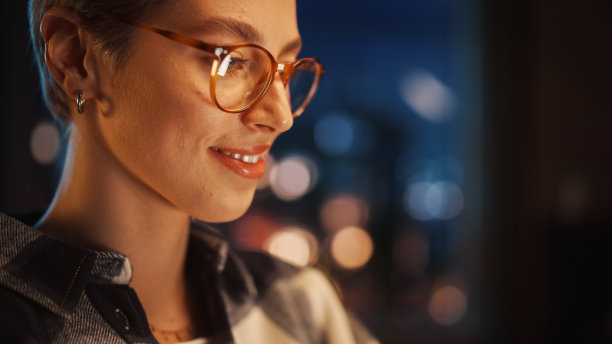 近距离肖像年轻美丽的女人使用电脑在时尚的阁楼公寓。有创意的女性与短头发和眼镜微笑检查社交媒体，浏览互联网。图片下载