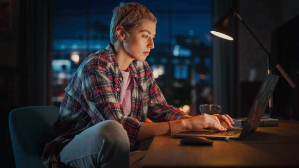 年轻迷人的女人使用笔记本电脑在时尚的阁楼公寓在晚上。创意女性微笑，检查社交媒体，浏览互联网。从大窗户看城市。图片下载