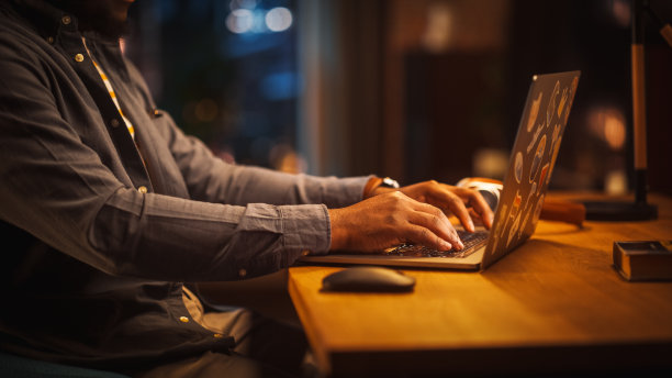 坐在桌子旁使用电脑的人的黑手的特写镜头。穿着灰色衬衫的创意设计师使用鼠标，在键盘上打字。显示器和笔记本电脑在桌上。图片下载