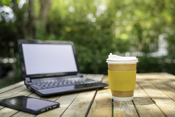 咖啡杯纸和笔记本电脑在绿色的自然背景。远程或在家工作。软的焦点。浅聚焦效果。图片下载