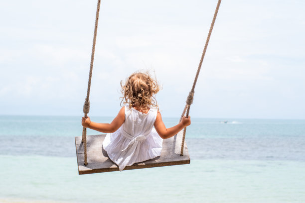 一个穿着白裙子的卷发小女孩在沙滩上的秋千上荡秋千。和孩子一起旅行和度假图片下载