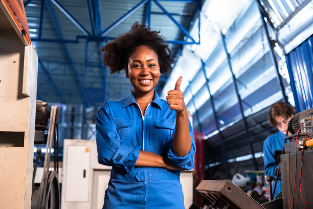 非裔美国人身穿防护服的年轻女工在工厂操作机器。在工业中工作的人。女工程师在工作场所看着相机的肖像。图片下载