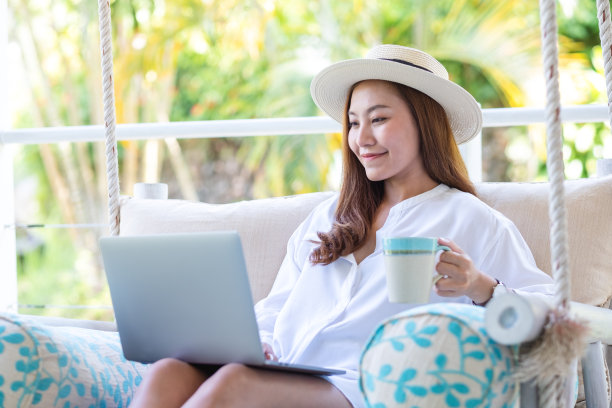 一个美丽的年轻亚洲女人使用笔记本电脑远程工作或在线学习的肖像形象，同时喝咖啡，坐在秋千沙发上图片下载