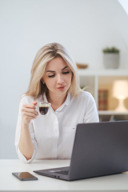 年轻漂亮的白种金发女人在家里用笔记本电脑工作，喝咖啡。远程工作或自由职业。图片下载