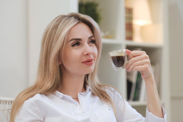 一位年轻美丽的白种人金发女子在家里休息。一个女人坐在桌旁，喝着杯子里的咖啡，读着杂志。图片下载