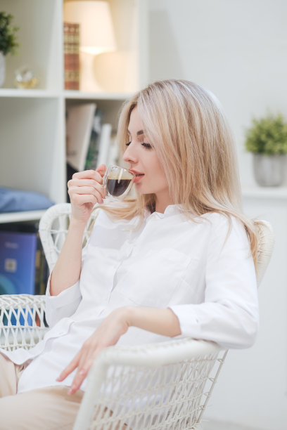 一位年轻美丽的白种人金发女子在家里休息。一个女人坐在桌旁，喝着杯子里的咖啡，读着杂志。图片下载