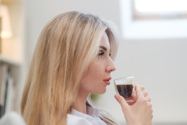 一位年轻美丽的白种人金发女子在家里休息。一个女人坐在桌旁，喝着杯子里的咖啡，读着杂志。图片下载