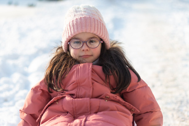 穿着冬季服装、针织帽子和眼镜的小孩坐在雪橇上向前看。户外冬季照片图片下载