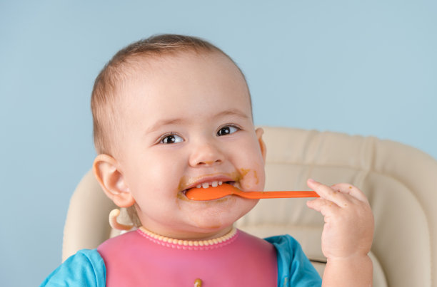 一名12-17个月、带着粉色围兜、面带微笑的新生儿把勺子放进嘴里，用牙齿咬着图片下载