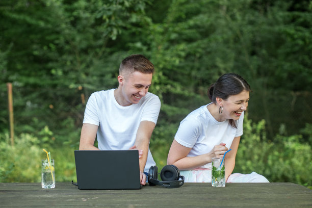 迷人的微笑青年男女坐在乡间别墅的院子里聊天。一起在笔记本电脑上工作。自由职业者使用技术，做笔记。冷却鸡尾酒和鸡尾酒。图片下载