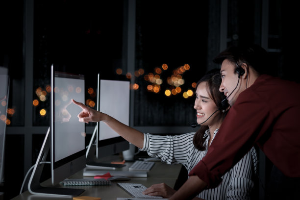 快乐的亚洲男女在呼叫中心咨询台与队友讨论，客服主管团队使用麦克风耳机在深夜办公室支持客户图片下载