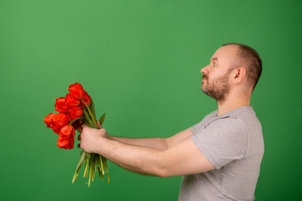一个没有刮胡子的中年男子在绿色背景下，把一束红色郁金香举到一旁。庆祝,道歉的概念。图片下载