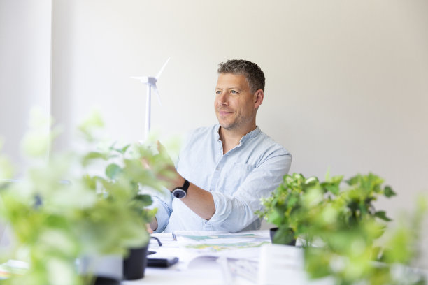 商业肖像的环境工程师坐在他的工作桌上，手里拿着一个风力涡轮机模型图片下载