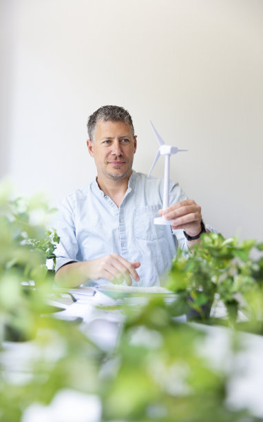商业肖像的环境工程师坐在他的工作桌上，手里拿着一个风力涡轮机模型图片下载