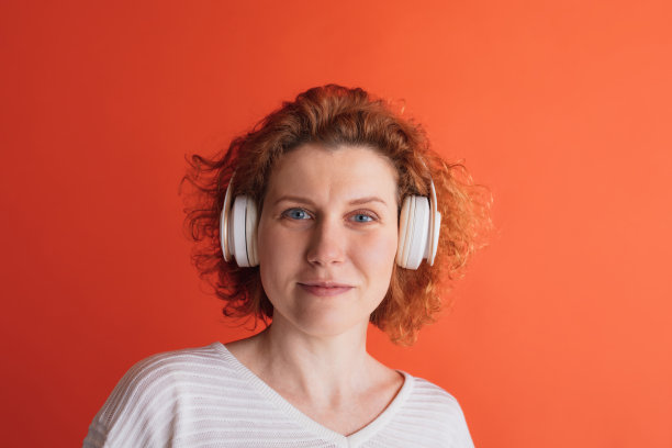 肖像美丽的红头发的女人明亮的蓝色眼睛听音乐在耳机和看着相机在红色的工作室背景图片下载