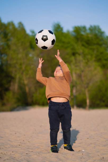 小孩用手把球抛起来。有趣的小男孩在沙滩上的公园里玩黑白足球图片下载