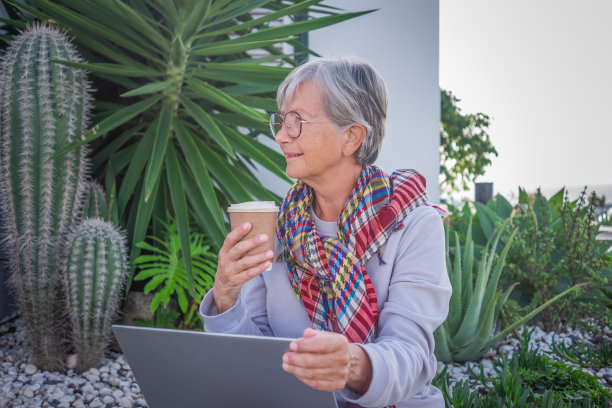 白发苍苍、成熟的老年女性在家外面用笔记本电脑坐在花园里，手里拿着咖啡杯。积极的老年人，无忧无虑的人，享受科技和社交。旅行或退休图片下载