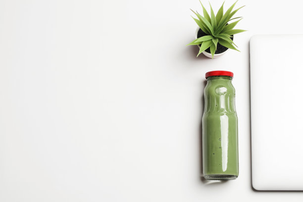 新鲜的绿色奶昔桌面上笔记本电脑和白色背景上的花。办公室里的健康零食。副本的空间。健康的生活方式的概念。图片下载