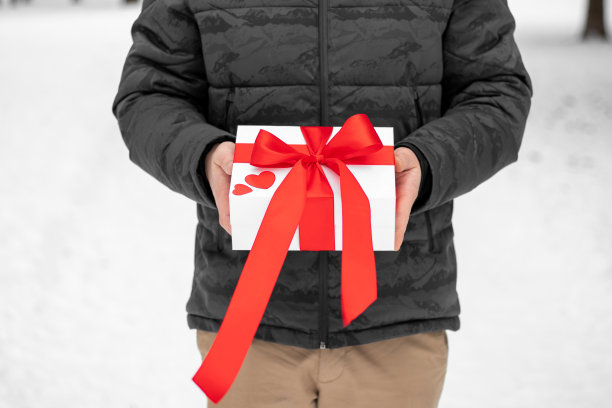 情人节礼物。一个男人手中的精美情人节礼品盒。男人为他的女朋友带来情人节礼物。带有红色丝带、蝴蝶结和心形的白色礼物。图片下载
