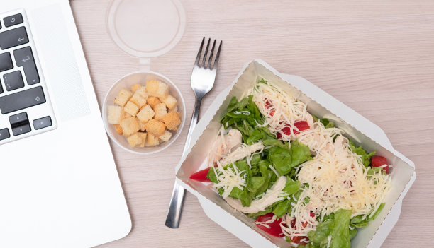 凯撒沙拉和鸡肉外卖包装，叉子和笔记本电脑在办公室的桌子上。食品递送和外卖食品概念图片下载