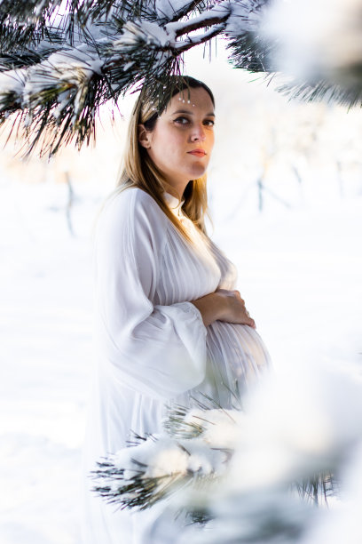 一个年轻漂亮的孕妇穿着白色的裙子站在雪地里，阳光照在她的脸上。怀孕。股票的照片。图片下载