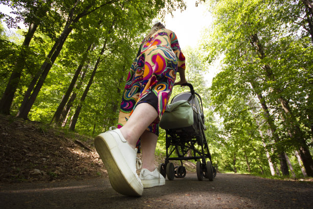 在公园里，一个穿着裙子和运动鞋的女人用婴儿车带着孩子走在后面的广角镜头。人们在自然界中。股票的照片。图片下载