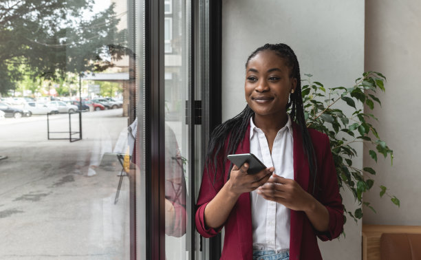 年轻的非洲裔美国女商人，穿着便服，站在餐馆的窗口附近，手里拿着手机，等着服务员把午餐装进盒子里。等待外卖咖啡的自由职业者图片下载