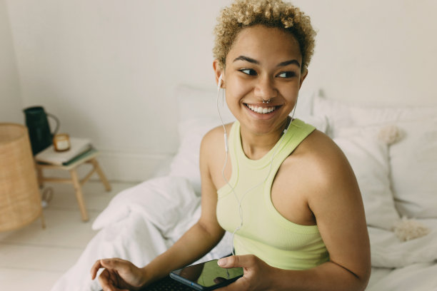 可爱的非洲女人坐在卧室的床上。快乐的女性在现代智能手机上听音乐，微笑着，高兴地看着别处。休闲时间和在线娱乐概念图片下载